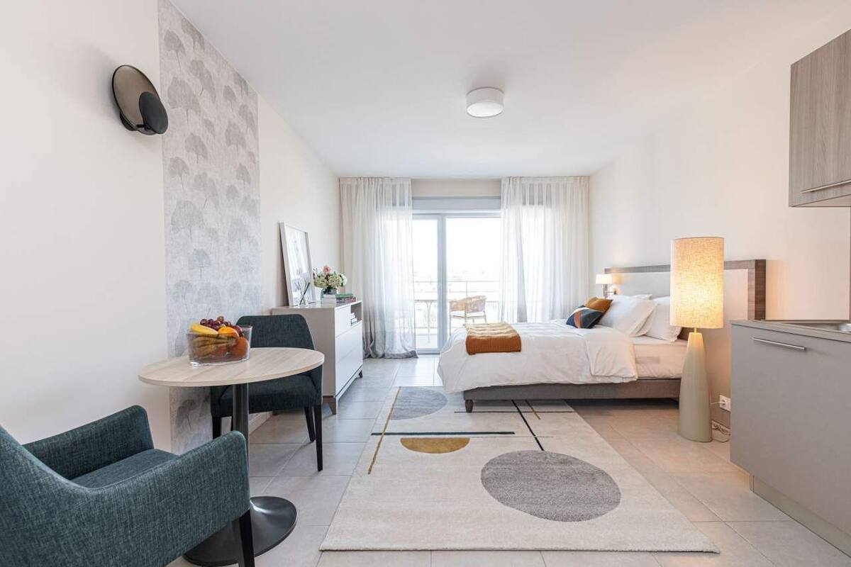 A modern hotel room with a bed, a desk, a chair, a lamp, and a view of the outdoors.