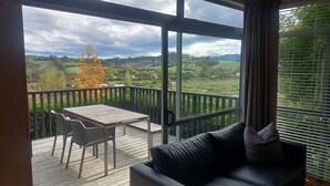 A balcony with a table and chairs, a sofa, and a view of a green landscape with trees and hills. - Riverside Matakana (Matakana)
