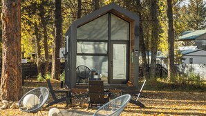 Eine Holzhütte mit großem Fenster, einem Sessel und einem Tisch in einem bewaldeten Gebiet.