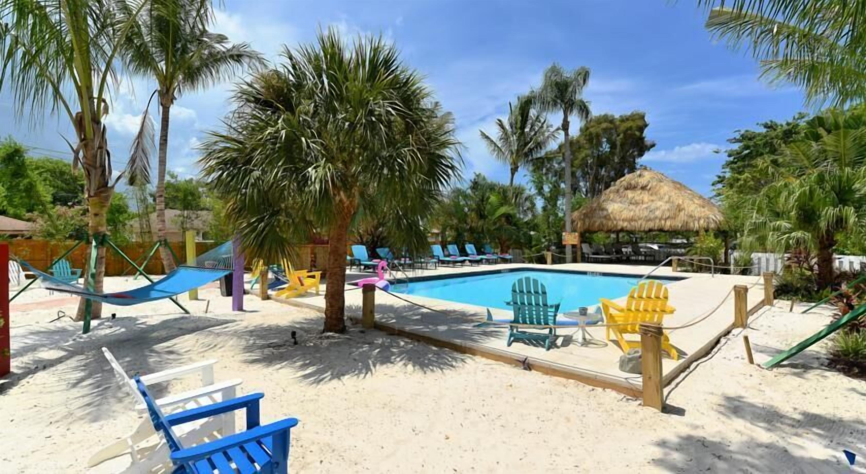 Un espace piscine agrémenté d’une structure à toit de chaume, entouré de palmiers et de chaises de plage.