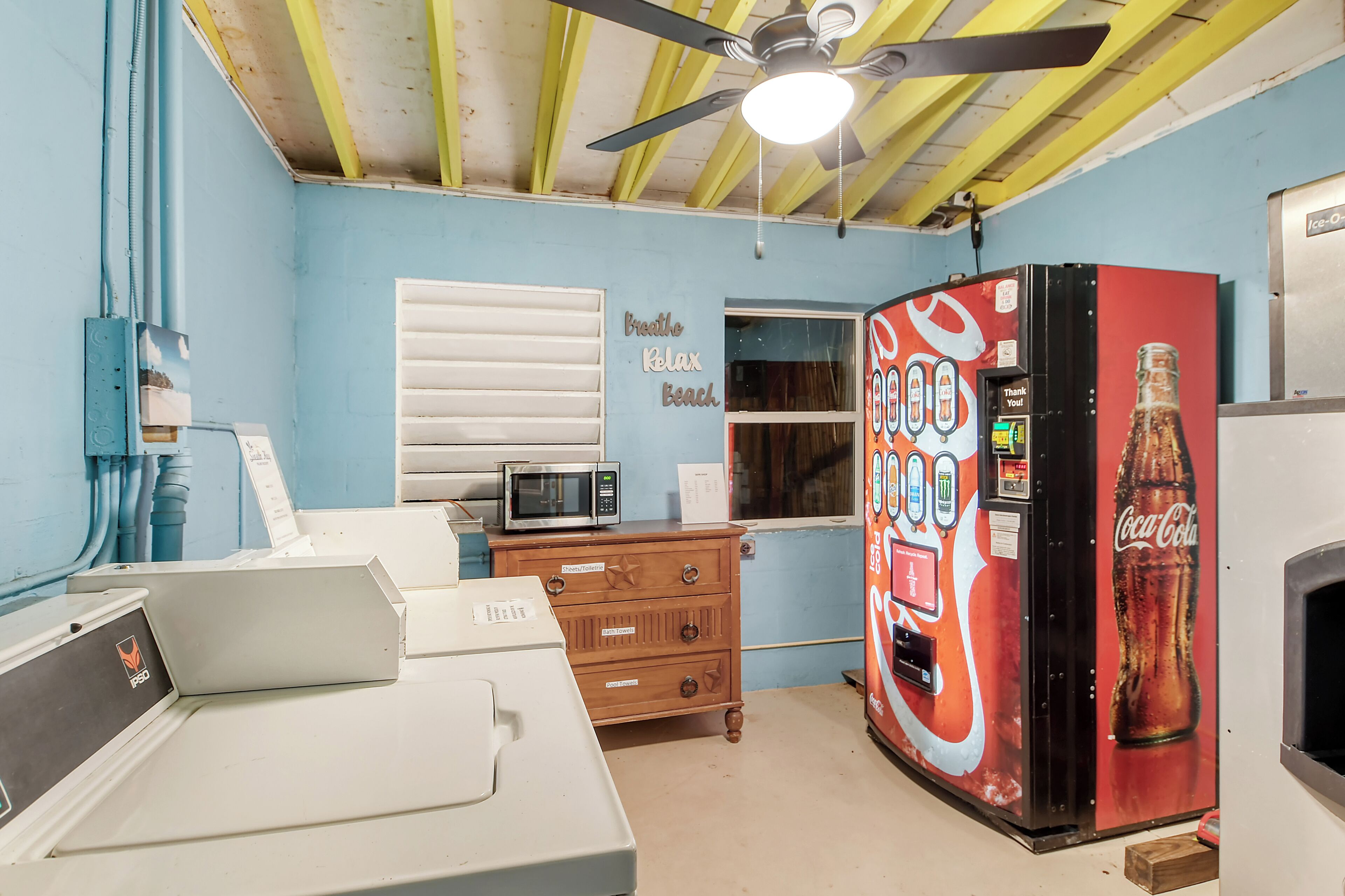 A room with a ceiling fan, a microwave, a Coca-Cola vending machine, a wooden dresser, and a window with blinds.
