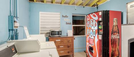 A room with a ceiling fan, a microwave, a Coca-Cola vending machine, a wooden dresser, and a window with blinds.