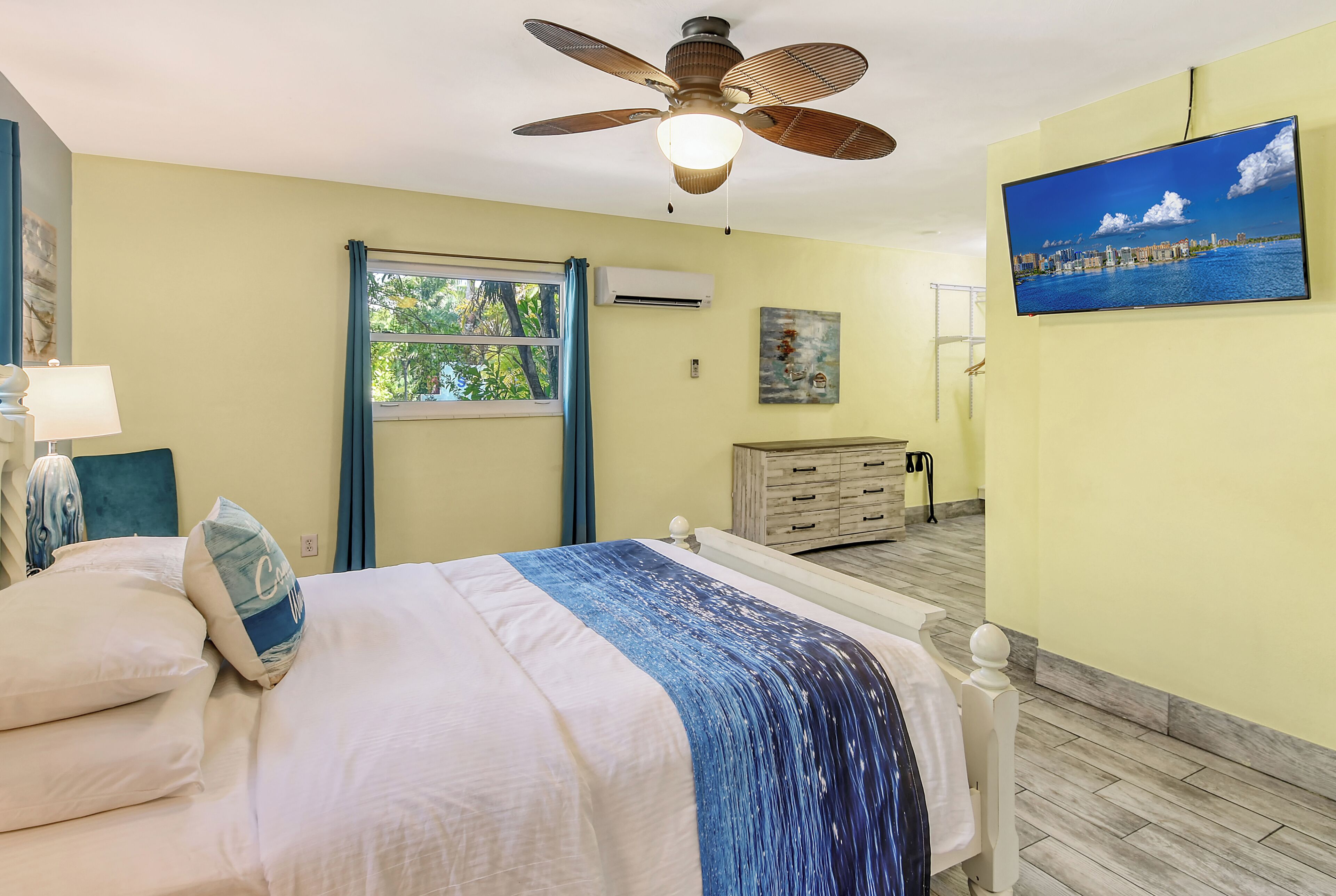 Une chambre à coucher avec un lit, un ventilateur de plafond, un appareil de climatisation, une fenêtre avec des rideaux, un tableau et une vue sur le paysage urbain par la fenêtre.