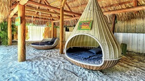 Un cabanon de plage doté d’un hamac et d’une chaise longue, entouré de sable et de structures en bois.