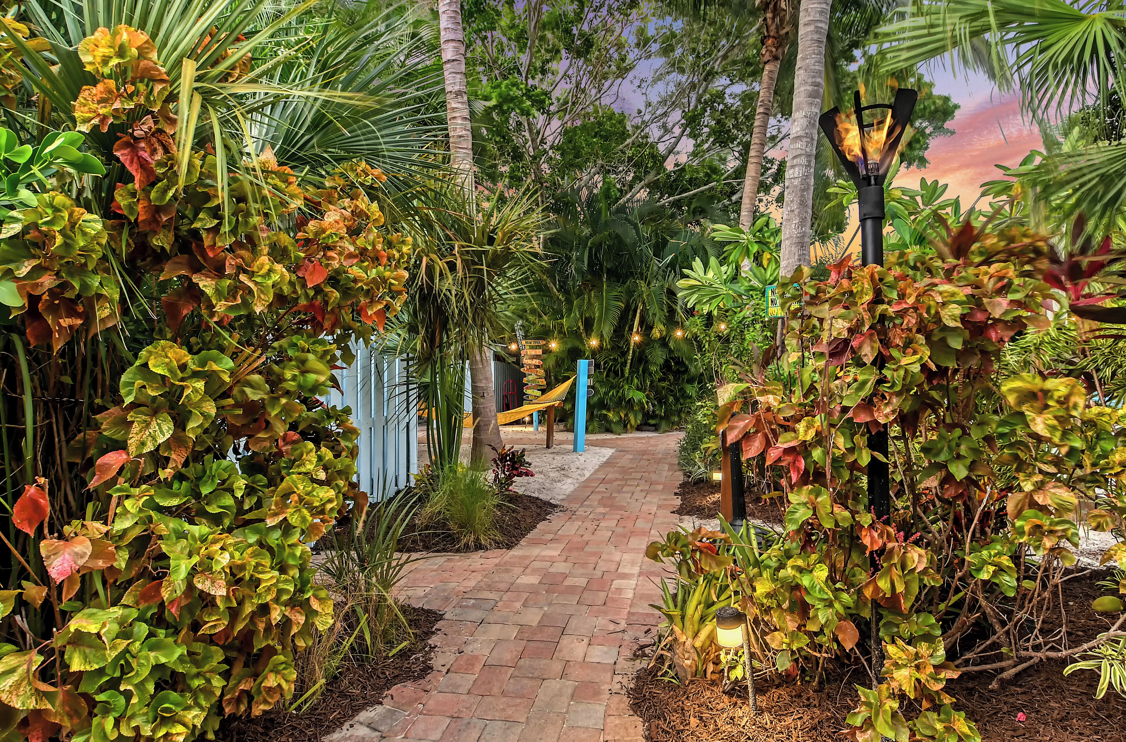 A garden pathway with a brick walkway, surrounded by lush greenery and palm trees.