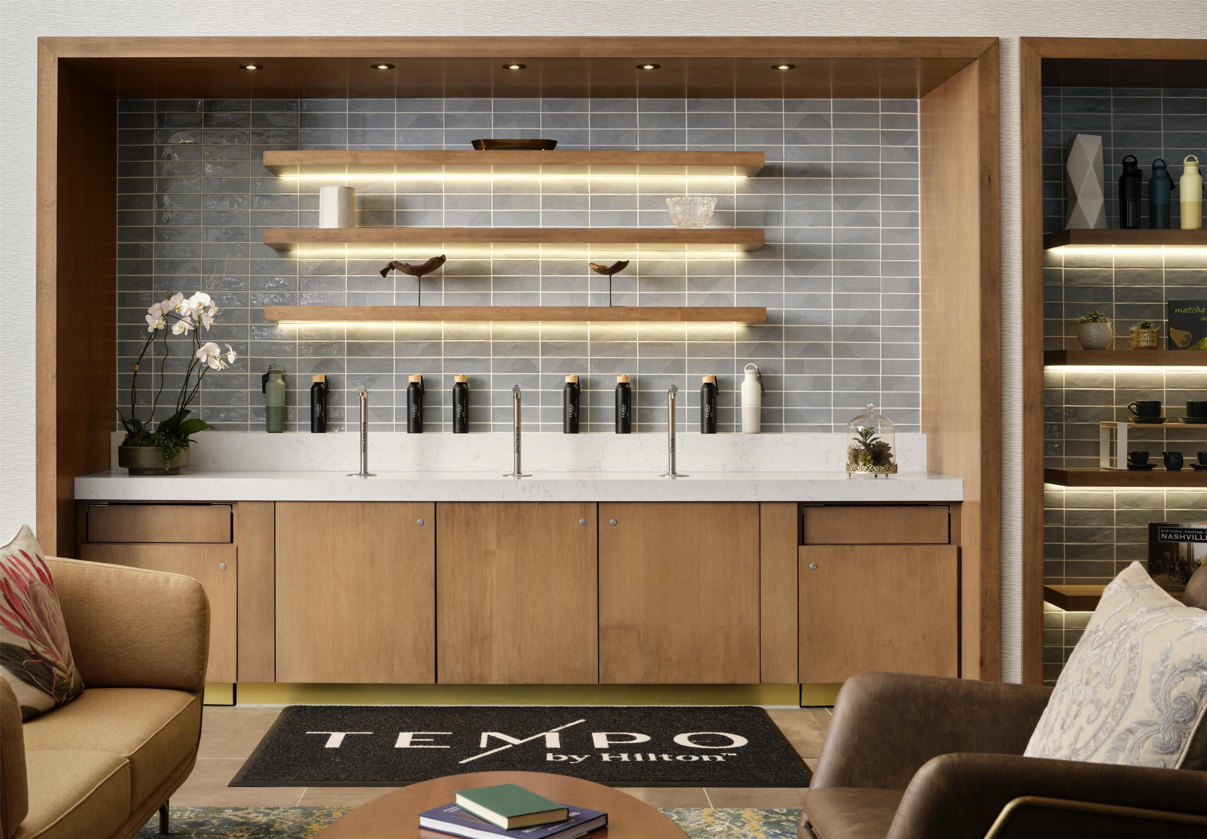 A modern kitchen with wooden cabinets, a white countertop, and a backsplash with blue tiles. There are shelves with kitchenware and a potted plant.