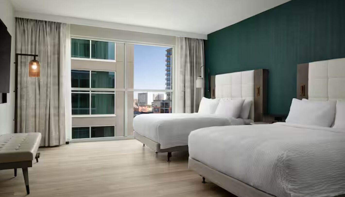 A hotel room with two beds, a bench, a large window with a view of buildings, and a green accent wall.