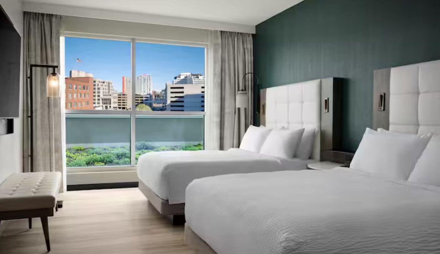 A hotel room with two beds, a bench, a large window with a city view, and a dark green accent wall.