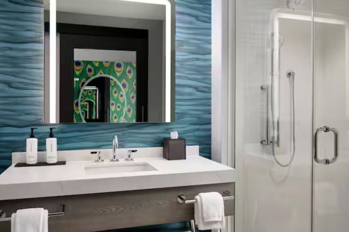 A modern bathroom with a peacock-themed mirror, a white sink with two bottles, and a glass shower enclosure.