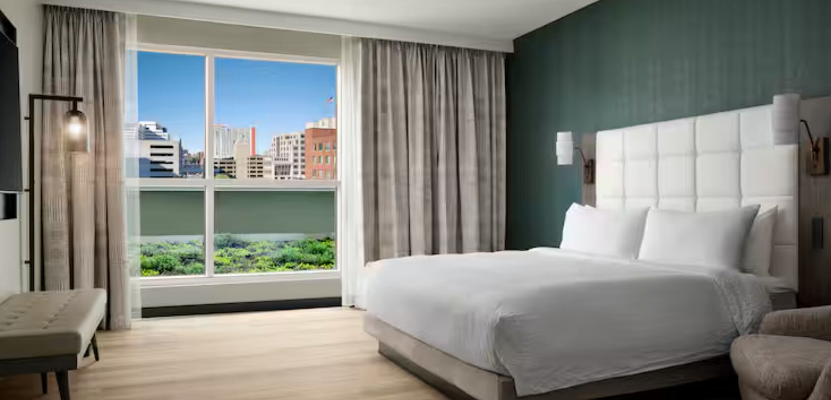 A hotel room with a large bed, a bench, a city view through the window, and a green wall.