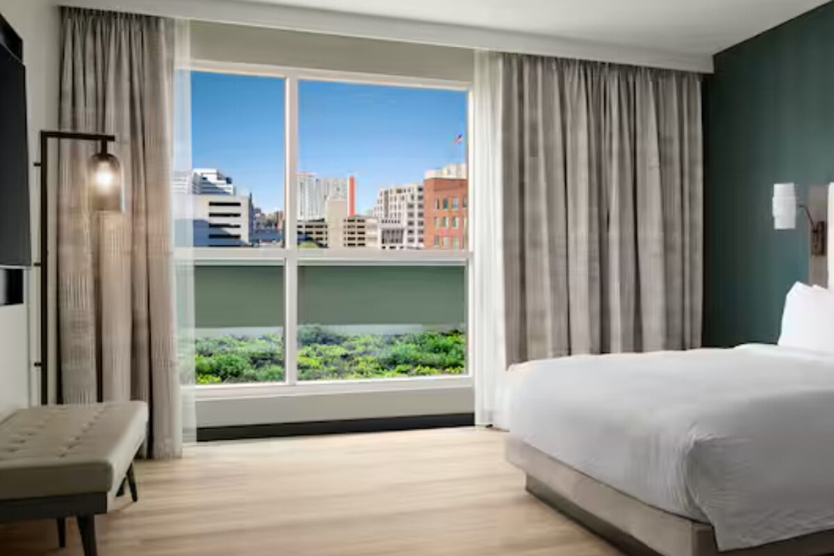 A hotel room with a large bed, a bench, a city view through the window, and a green wall.