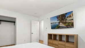 Et soveværelse med en stor seng, et træskab, en hvid dør og et billede af et kystlandskab hængt på væggen.
