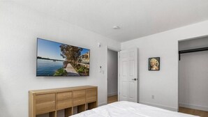 Une chambre à coucher avec un grand lit, une commode en bois, un tableau représentant un paysage côtier et une porte donnant sur une autre pièce.