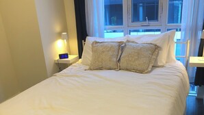 Ein ordentlich bezogenes Bett mit weißen Leinen und zwei Zierkissen, ein Nachttisch mit Lampe sowie ein Fenster mit Vorhängen.