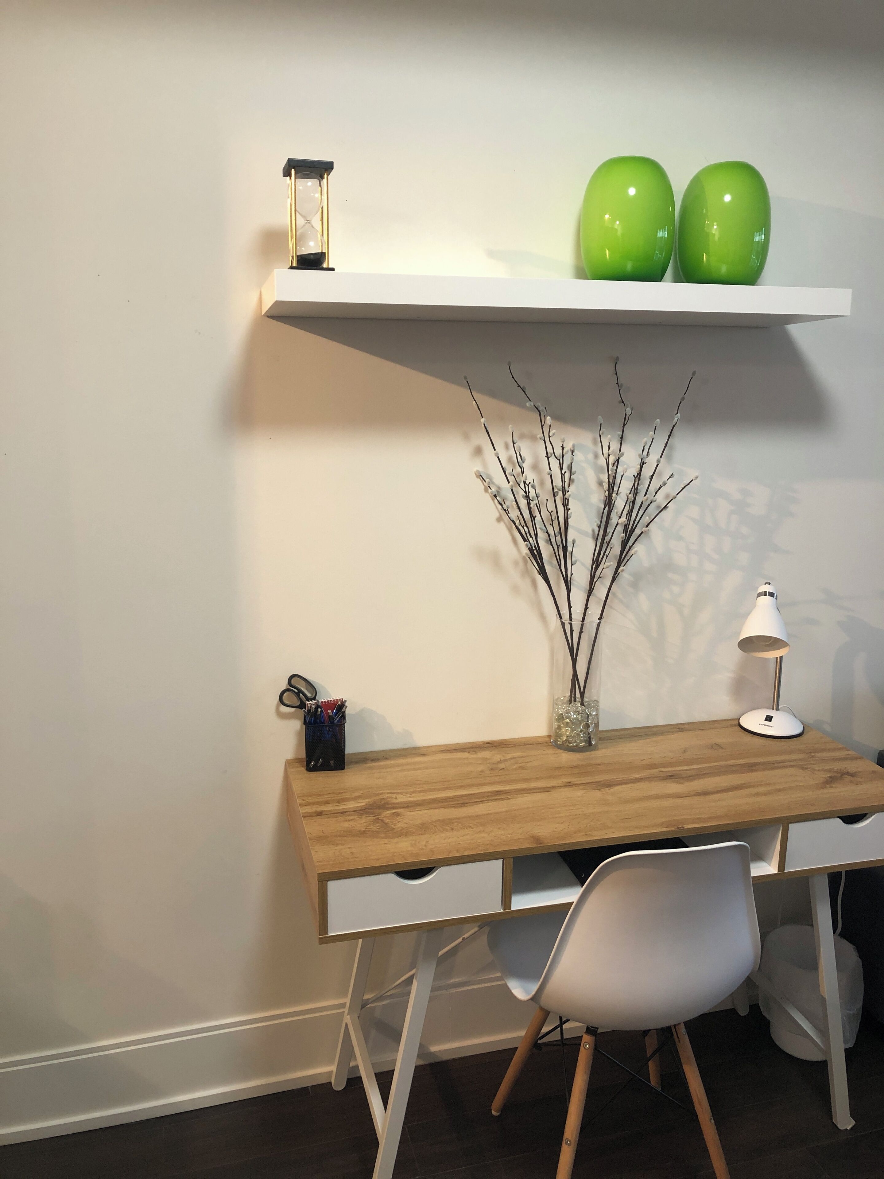 A wooden desk with a white chair, a small shelf with two green objects, a lamp, and a vase with branches.