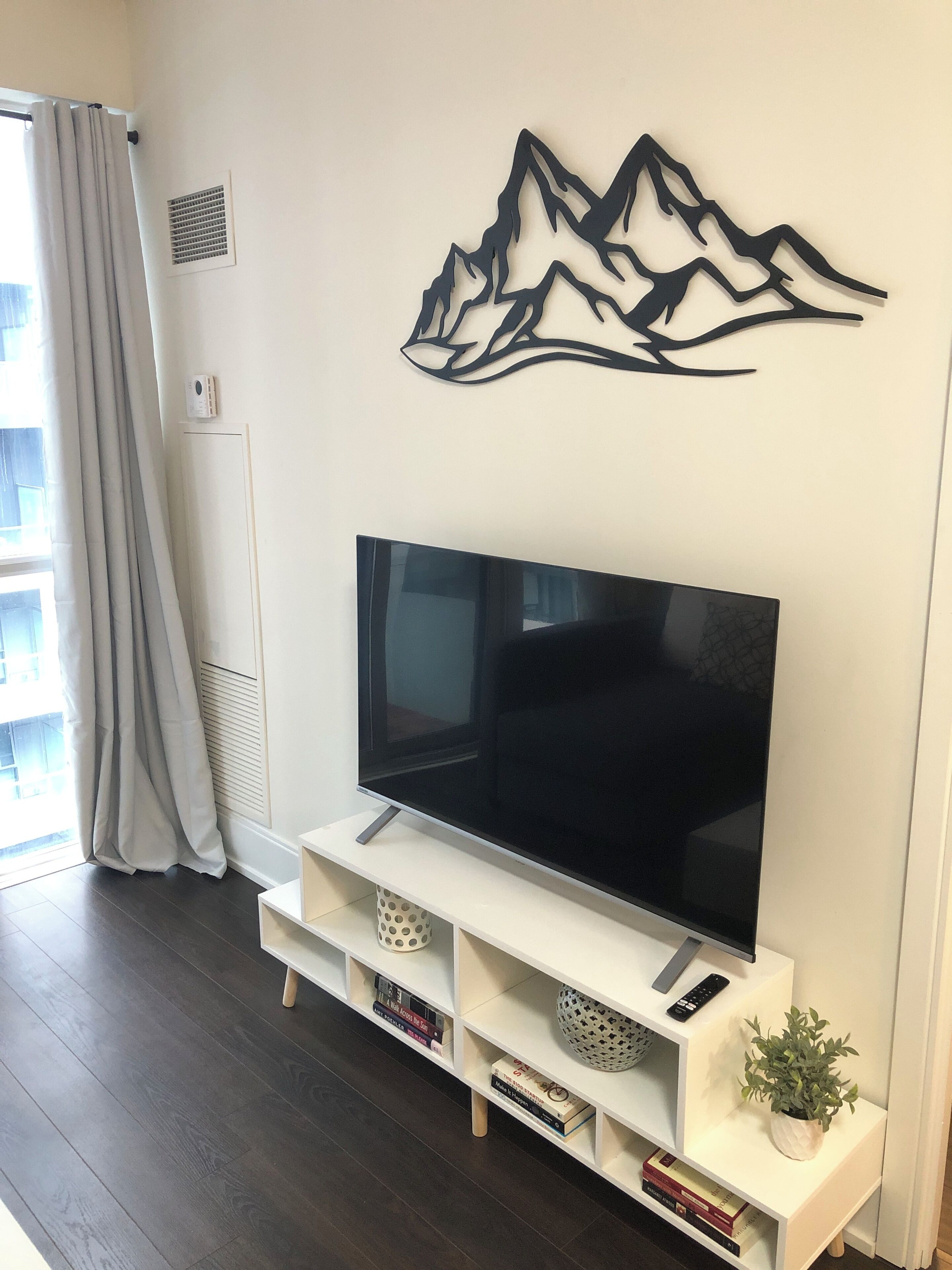 A modern living room with a flat-screen TV on a white stand, a decorative mountain wall art piece, and a potted plant on the stand.