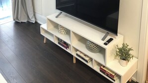 Ein modernes Wohnzimmer mit einem Flachbildfernseher auf einem weißen Lowboard, einem dekorativen Wandbild mit Bergmotiven und einer Blumentopf-Pflanze auf dem Lowboard.