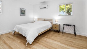 A bedroom with a wooden bed, a nightstand, a small table, a window with blinds, and an air conditioning unit. - Designer Loft in Pueblo – Central Modern Comfort (Pueblo)