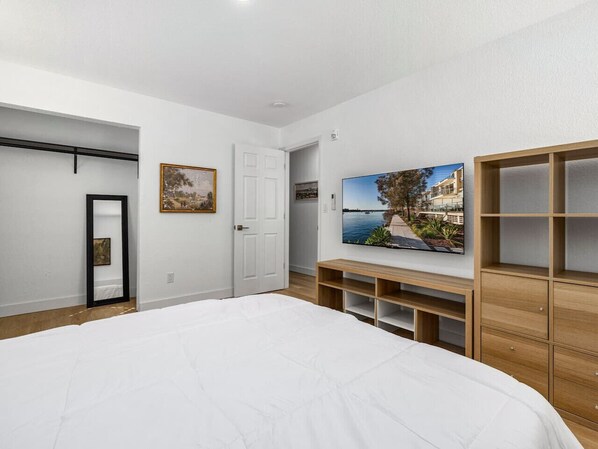A bedroom with a large bed, a wooden wardrobe, a TV mounted on the wall, and a door leading to another room.