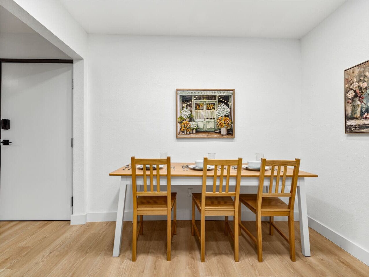 Un coin repas avec une table blanche et quatre chaises en bois. Un tableau encadré est accroché au mur, au-dessus de la table.