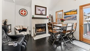 A living room with a fireplace, a dining table with chairs, a sofa, and a chair. There are paintings on the wall and a sign with a ski patrol logo. - 3B/2BA + Loft | Eagle-Vail• Dog-Friendly (Avon)
