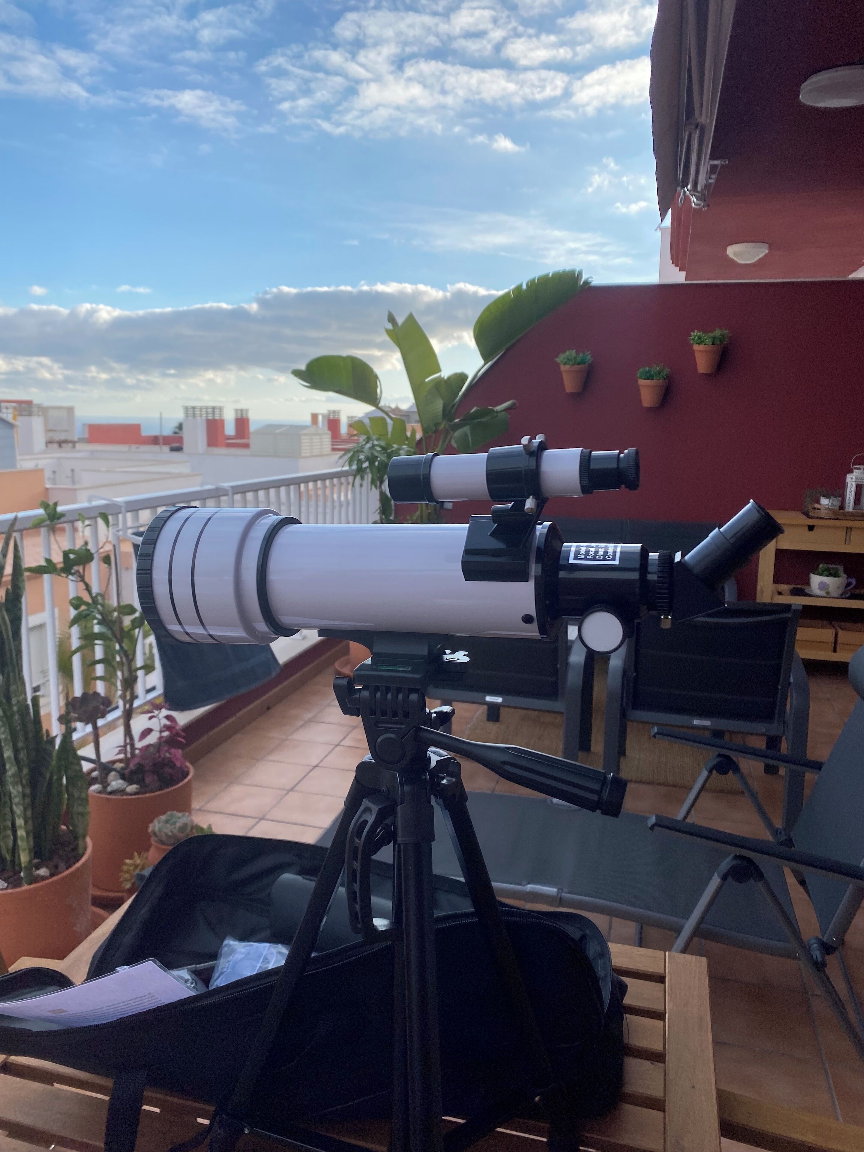 A telescope on a balcony with potted plants and a red wall in the background.