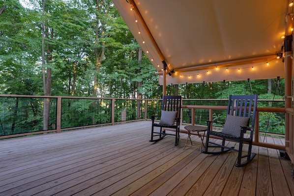 Um deck de madeira com duas cadeiras de balanço e uma mesa, rodeado por um toldo branco e luzes de Natal.