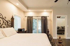 A bedroom with a large bed, a bedside table with a radio, a window with blinds, and a view of a balcony and another room.