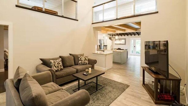 Un ampio soggiorno con un grande divano, un tavolino da caffè e una TV su un mobile in legno. Si intravede una cucina sullo sfondo, con soffitto alto e ampie finestre.