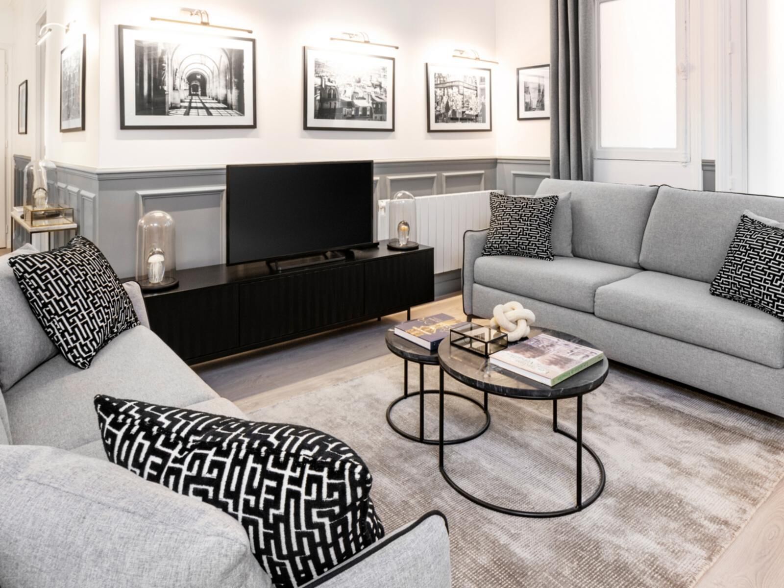 En moderne stue med en grå sofa, puder med sort og hvide mønstre, et sort multimediemøbel og et fladskærms-tv.