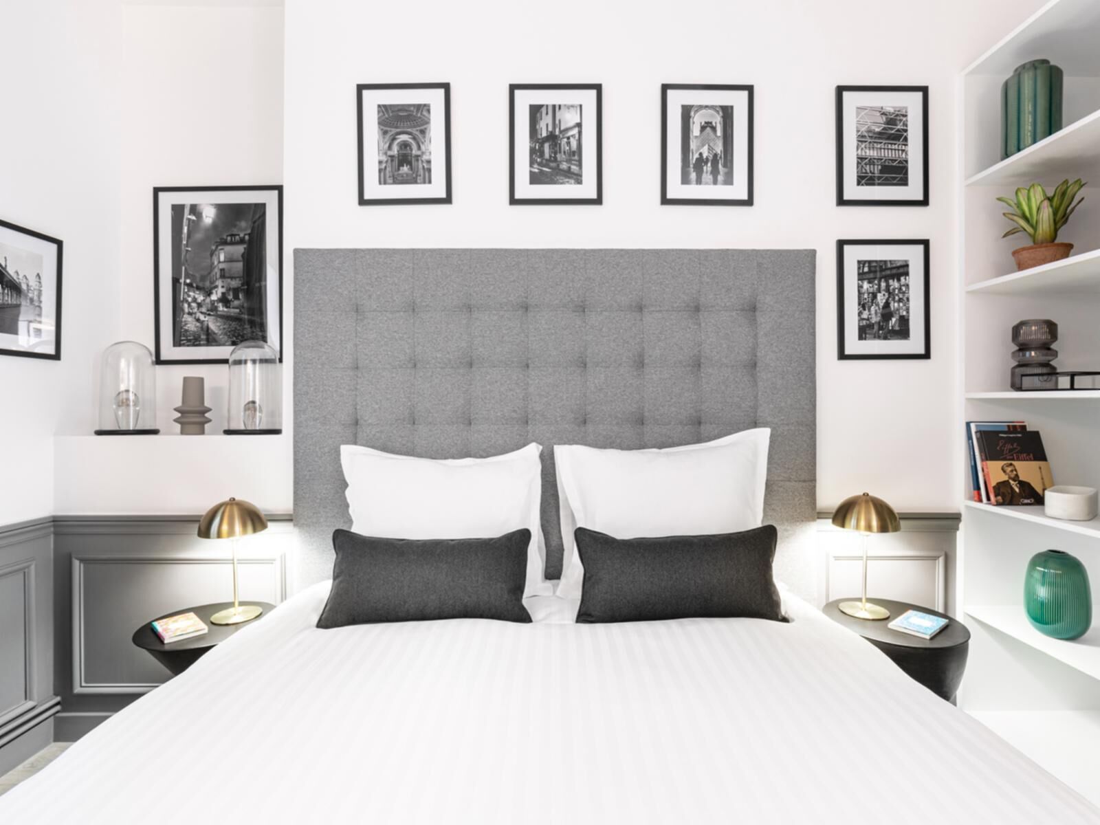 Een moderne slaapkamer met een gestoffeerd hoofdeinde, twee witte kussens, een zwart kussen, een nachtkastje met een lamp en een boekenplank met boeken en decoratieve objecten.