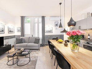 Un salon moderne avec un canapé gris, des chaises noires, une table à manger en bois ornée d’un vase de fleurs, et une cuisine en arrière-plan.
