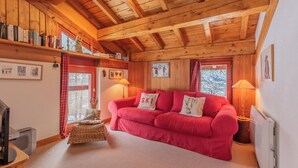 Ein gemütliches Zimmer mit einem roten Sofa, Holzbalkendecke und einem Fernseher auf einem Holztisch.