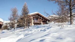 Eine Holzhütte mit verschneitem Dach, umgeben von Bäumen und verschneiter Landschaft.