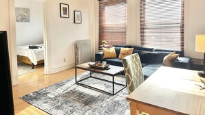 Ein modernes Wohnzimmer mit einer Couch, einem Couchtisch und einem Esstisch. Zwei Fenster mit Jalousien, eine Heizung und ein Bett im angrenzenden Zimmer.