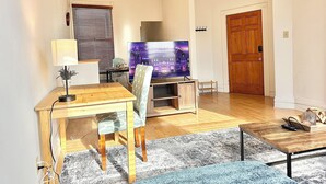 Ein Wohnzimmer mit einem Holzesstisch, einem Fernseher auf einem Schrank, einem blauen Sofa und einer Holztür.