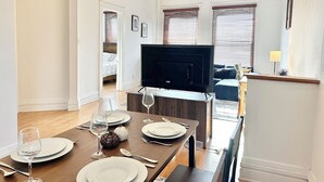 Ein modernes Wohnzimmer mit Flachbildfernseher, einem Esstisch für zwei Personen und einer gemütlichen Sitzecke mit Sofa und kleinem Tisch.