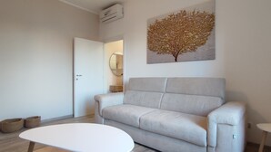Un salon moderne avec un canapé beige, une table basse blanche et une petite table d’appoint. Un tableau représentant un arbre est accroché au mur, et une porte donne accès à une autre pièce.