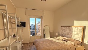 Une chambre à coucher comprenant un lit, un bureau, une armoire et une fenêtre donnant sur des immeubles.