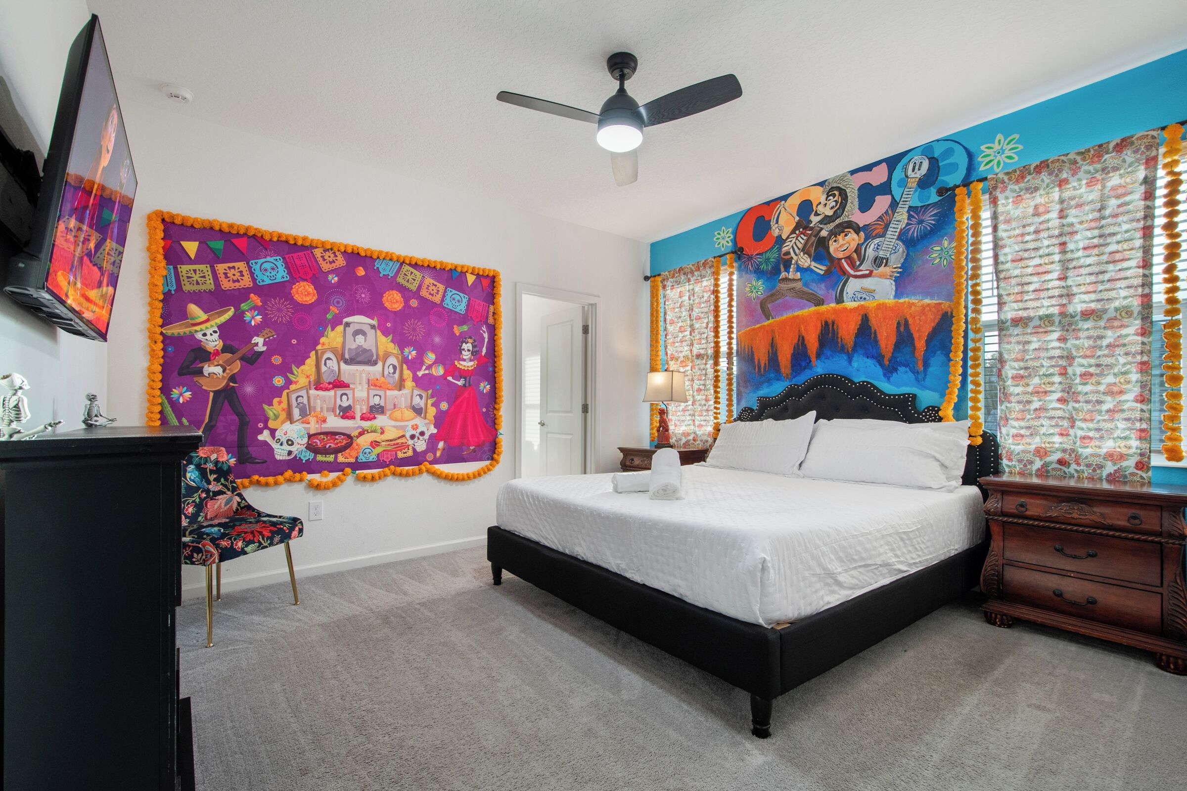 Un dormitorio con cama, mesita de noche, silla, ventilador de techo y televisor en la pared.