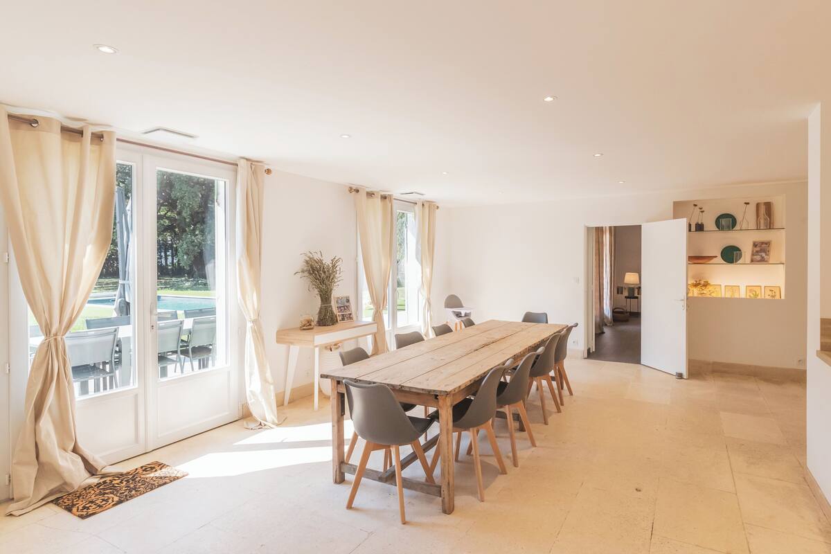 Een ruime eethoek met een houten tafel en stoelen, grote ramen met gordijnen en uitzicht op de omgeving.