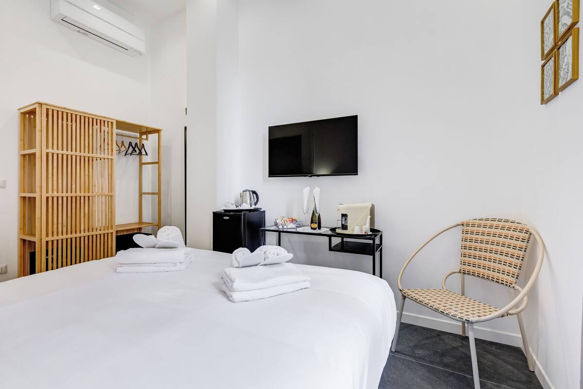 Une chambre d’hôtel avec un grand lit, une armoire en bois, un bureau avec un téléviseur à écran plat, un fauteuil en osier et une petite table avec des articles de toilette.