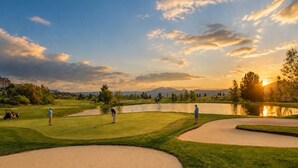 Een golfbaan met zandbunkers, een vijver en een ondergaande zon op de achtergrond.