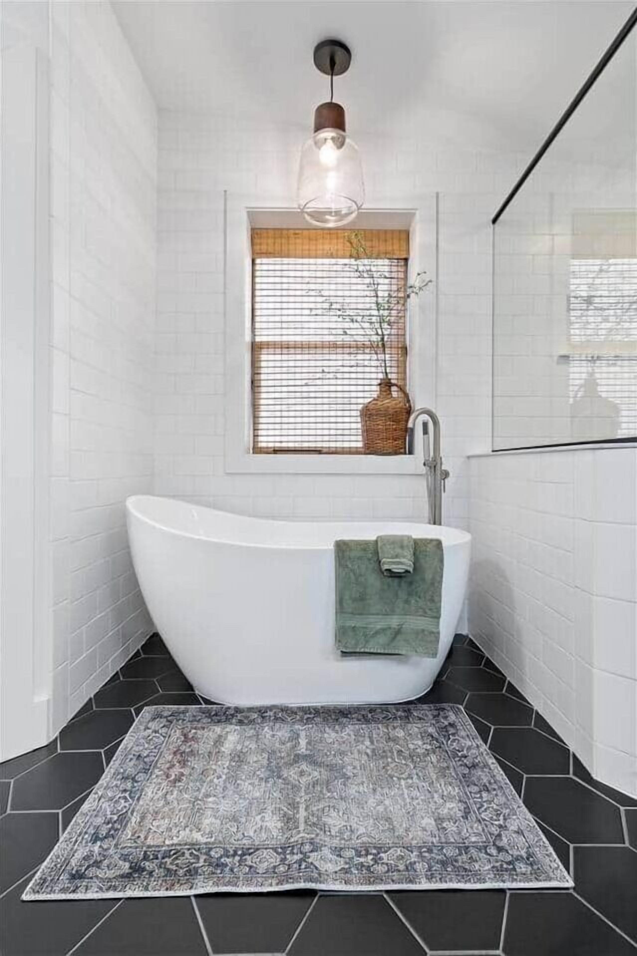 A modern bathroom with a freestanding bathtub, a window with a wicker basket, and a patterned rug on the floor.