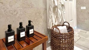 A bathroom with a wooden table holding three bottles labeled "Body Wash," "Shampoo," and "Conditioner." There is a wicker basket with a towel on a white mat. - Veranera 1 | Relaxing Private Heated Pool at Aguadilla (Aguadilla)