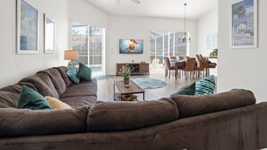 A spacious living room with a brown sofa, a coffee table, and a TV mounted on the wall. There is a dining area with chairs and a table, and a ceiling fan. - 293 - Fabulous 3 Bed Villa with Private Oversized Pool (Davenport)