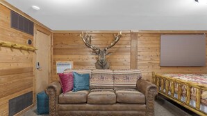A log cabin room with a brown leather sofa, two pillows, a mounted deer head, a wooden bed frame, and a window with blinds.