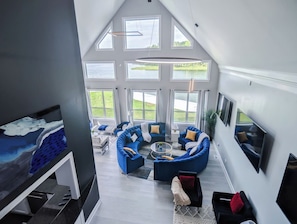 A spacious living room with a large window, a curved blue sofa set, and a flat-screen TV mounted on the wall.