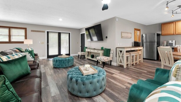 A modern living room with a sofa set, ottomans, a flat-screen TV, a kitchen area, and a dining table.