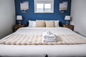 A neatly made bed with a fluffy white blanket and a rolled towel, two bedside lamps, and a window with a view of the sky.